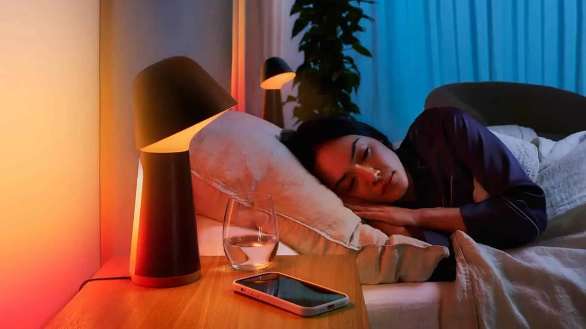 Cảnh một người phụ nữ đang ngủ say trên giường, cạnh đó là điện thoại thông minh và đèn ngủ thông minh, cho thấy ứng dụng tiếng ồn trắng hỗ trợ giấc ngủ.
