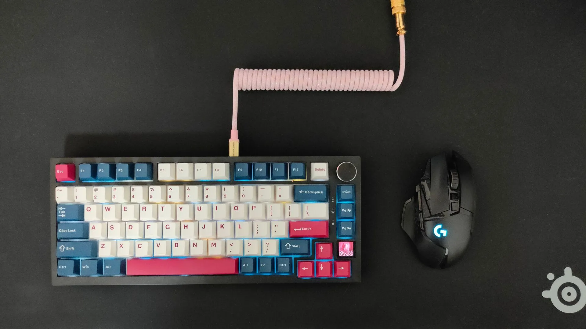 Một bàn phím cơ với dây cáp xoắn màu hồng và chuột không dây Logitech G502 trên tấm lót chuột SteelSeries QcK Performance màu đen.
