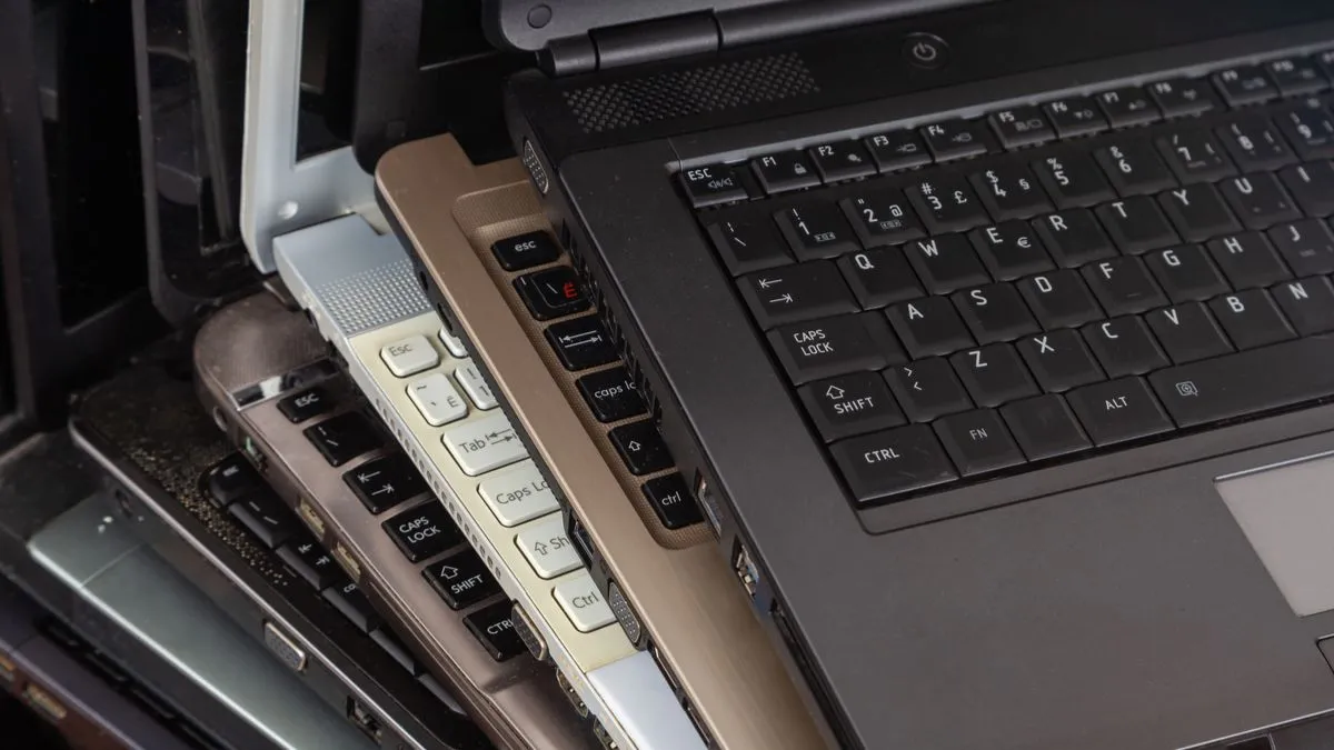 Một chồng các laptop cũ bám bụi với nhiều màu sắc và mẫu mã khác nhau, gợi lên hình ảnh về công nghệ đã qua sử dụng.